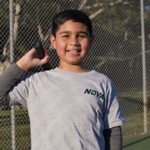 Boy in a NOVA T-shirt holding up a tennis ball by the court fence